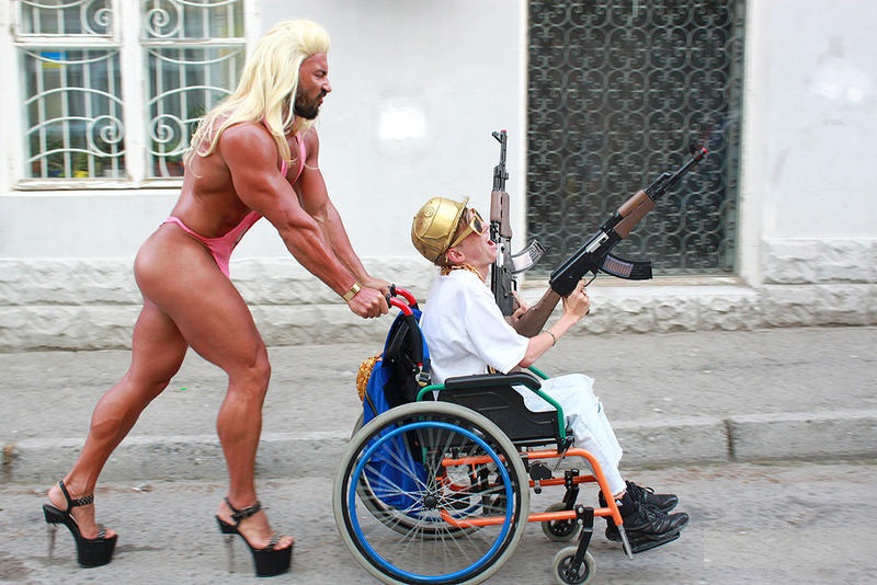 Photo d'un body builder nu, travesti, poussant un homme en chaise roulante armé de mitrailleuses